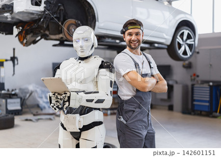 A smiling mechanic stands with a humanoid robot in a modern auto repair garage with a car lifted for service 142601151