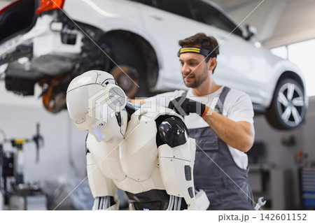 A skilled technician in overalls and gloves is carefully repairing a humanoid robot in a brightly lit automotive workshop A skilled technician in overalls and gloves is carefully repairing a humanoid robot in a brightly lit automotive workshop 142601152