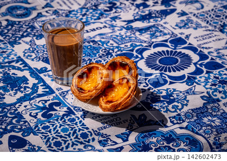 Three quarter view of Portuguese custard tarts and glass of meia de leite coffee on blue and white tablecloth in warm outdoor light and blurred background. Famous local dessert, cultural food, travel Three quarter view of Portuguese custard tarts and glass of meia de leite coffee on blue and white tablecloth in warm outdoor light and blurred background. Famous local dessert, cultural food, travel 142602473