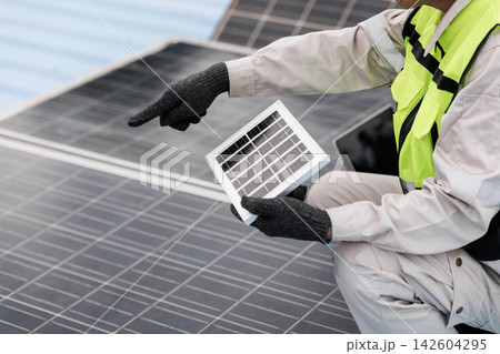 Solar energy technician in safety gear holds a small solar panel and points at a rooftop array during inspection or maintenance. 142604295