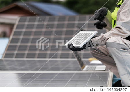 Solar energy technician in safety clothing holds a small solar panel while working beside rooftop photovoltaic panels outdoors. Solar energy technician in safety clothing holds a small solar panel while working beside rooftop photovoltaic panels outdoors. 142604297
