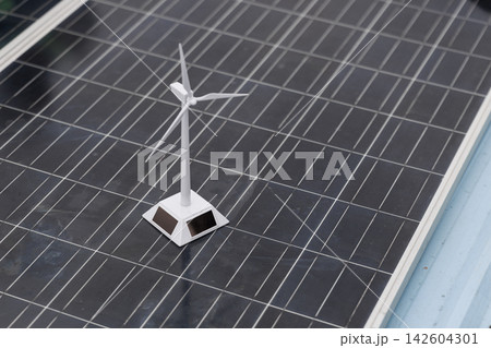 Small white wind turbine model standing on dark solar panels, clean energy and renewable power technology. Small white wind turbine model standing on dark solar panels, clean energy and renewable power technology. 142604301