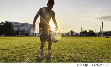 Young man juggling soccer ball on stadium at sunset. Professional footballer kicking ball at green field. Sportsman practicing tricks at meadow with sunlight at background. Freestyle football Young man juggling soccer ball on stadium at sunset. Professional footballer kicking ball at green field. Sportsman practicing tricks at meadow with sunlight at background. Freestyle football 142604628