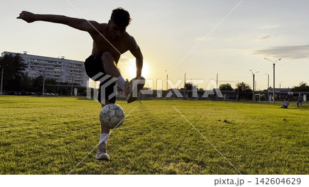 Young man juggling soccer ball on stadium at sunset. Professional footballer kicking ball at green field. Sportsman practicing tricks at meadow with sunlight at background. Freestyle football Young man juggling soccer ball on stadium at sunset. Professional footballer kicking ball at green field. Sportsman practicing tricks at meadow with sunlight at background. Freestyle football 142604629