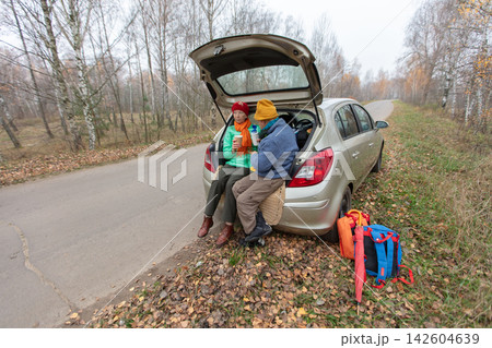 An elderly couple enjoys snacks while sitting by a parked car on the side of the road surrounded by trees in autumn. An elderly couple enjoys snacks while sitting by a parked car on the side of the road surrounded by trees in autumn. 142604639