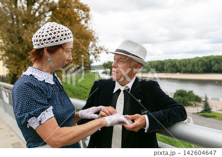 Elderly couple dressed in vintage clothing enjoying a moment by the river during a cloudy day Elderly couple dressed in vintage clothing enjoying a moment by the river during a cloudy day 142604647
