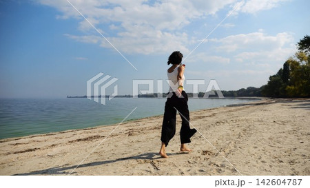 Happy adult woman jogging on sandy beach near shoreline at sunny day. Beautiful lady running on seashore with calm waves at background. Concept of summer vacation or holiday. Slow motion 142604787
