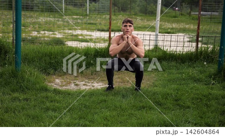 Male shirtless athlete doing squats at sports ground. Young sportsman working at summer nature. Strong sporty man exercising outdoor. Concept of active and sportive lifestyle. Slow motion Male shirtless athlete doing squats at sports ground. Young sportsman working at summer nature. Strong sporty man exercising outdoor. Concept of active and sportive lifestyle. Slow motion 142604864