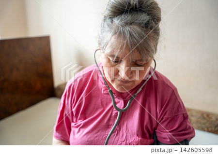 Mature woman in pink top checking her wellbeing with a stethoscope at home. Menopause health awareness selfcare and climacteric support 142605598