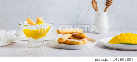 Mango trifle with savoiardi in a glass, sliced fruit, and cookies on plates in a web banner. 142605807