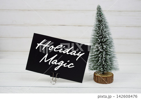 Holiday Magic text written on paper card with snowy tree on wooden background Holiday Magic text written on paper card with snowy tree on wooden background 142608476
