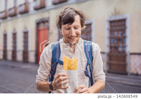 Man traveler holding a golden molote poblano traditional fried Mexican empanada outdoors on a street in Puebla Mexico with colonial city background paper wrapper local street food authentic travel. Man traveler holding a golden molote poblano traditional fried Mexican empanada outdoors on a street in Puebla Mexico with colonial city background paper wrapper local street food authentic travel. 142608549