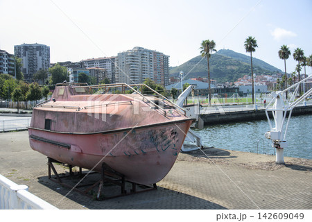 Weathered pink lifeboat on a cradle on the quay with the town and hill behind 142609049