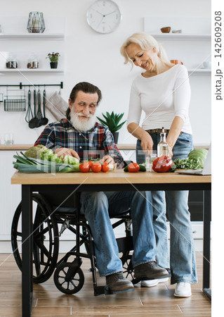 An elderly couple happily prepares a fresh vegetable meal in their kitchen, with the man in a wheelchair assisting An elderly couple happily prepares a fresh vegetable meal in their kitchen, with the man in a wheelchair assisting 142609208