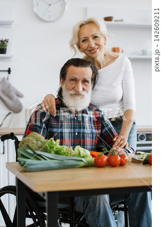 A smiling elderly couple, one in a wheelchair, preparing fresh vegetables in a bright kitchen, showcasing companionship and healthy living A smiling elderly couple, one in a wheelchair, preparing fresh vegetables in a bright kitchen, showcasing companionship and healthy living 142609211