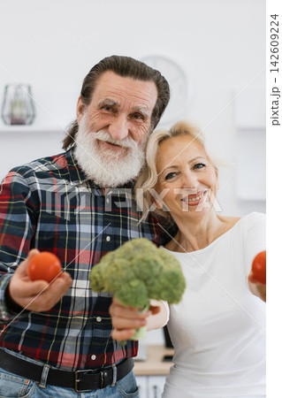 A joyful elderly couple smiles while holding fresh broccoli and tomatoes, embodying a healthy lifestyle and shared happiness in the kitchen A joyful elderly couple smiles while holding fresh broccoli and tomatoes, embodying a healthy lifestyle and shared happiness in the kitchen 142609224