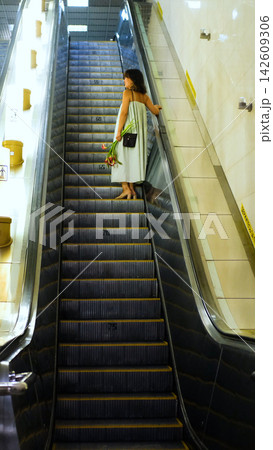A woman rides an escalator holding flowers; she wears a loose light-colored dress and a dark shoulder bag, set against a backdrop of long gray steps, cream-colored walls, and warm lighting in a subway 142609306