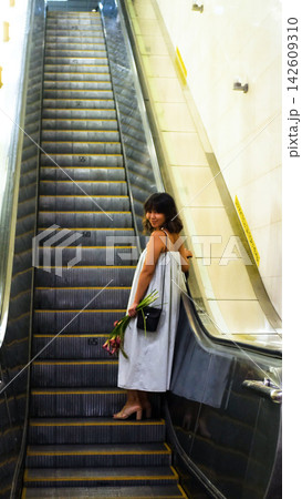 A woman rides an escalator holding flowers; she wears a loose light-colored dress and a dark shoulder bag, set against a backdrop of long gray steps, cream-colored walls, and warm lighting in a subway A woman rides an escalator holding flowers; she wears a loose light-colored dress and a dark shoulder bag, set against a backdrop of long gray steps, cream-colored walls, and warm lighting in a subway 142609310