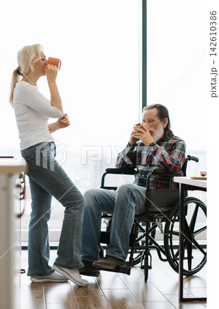 A senior couple shares a quiet moment drinking coffee together by a large window, one seated in a wheelchair A senior couple shares a quiet moment drinking coffee together by a large window, one seated in a wheelchair 142610386