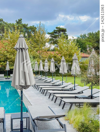 Resort Swimming Pool with Rows of Sun Loungers. Modern outdoor swimming pool with sun loungers, closed parasols, cloudy sky. Peaceful resort environment for relaxation, wellness, vacation travel 142611063