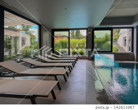 Indoor Swimming Pool with Spa Loungers. Modern indoor swimming pool with rows of relaxation loungers, large windows and garden views. Contemporary spa and wellness interior for leisure, relaxation Indoor Swimming Pool with Spa Loungers. Modern indoor swimming pool with rows of relaxation loungers, large windows and garden views. Contemporary spa and wellness interior for leisure, relaxation 142611067