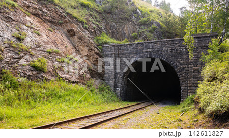 Old stone railway tunnel entrance built into a rocky hillside at Lake Baikal, Russia. Rusty train tracks leading into a dark archway surrounded by green summer trees 142611827