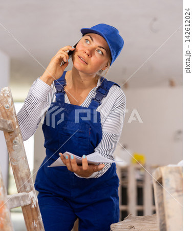 Female builder in blue overalls holding papers and talking on phone 142612024