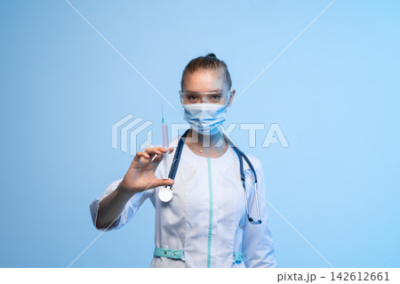 Healthcare professional in mask and goggles holding a syringe against a light blue background during vaccination and treatment campaign 142612661