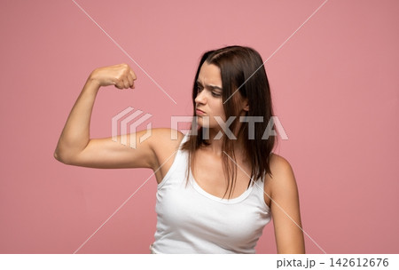 Young woman flexing biceps, showing determination and strength on a pink background, embracing power and confidence Young woman flexing biceps, showing determination and strength on a pink background, embracing power and confidence 142612676