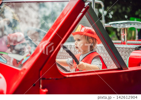 Little boy dressed as firefighter playing in a red toy fire truck at an outdoor playground with helmet safety vest pretend rescue game childhood dreams role play and playing actively. 142612904
