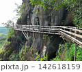 Wooden walkway around the steep sandstone cliff at Wat Phu Tok temple. Scenic mountain path trail with a wooden fence overlooking the green tropical forest and cloudy sky in Thailand 142618159