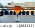 A glass of beer sits on a table outside a cafe during the day while cars pass by. 142623389