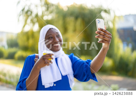 A smiling African American Muslim woman in a hijab holds a coffee cup while taking a selfie outdoors A smiling African American Muslim woman in a hijab holds a coffee cup while taking a selfie outdoors 142623888