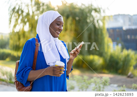 A happy African American Muslim woman wearing a white hijab and blue dress holds a coffee cup and smartphone while walking outdoors 142623893