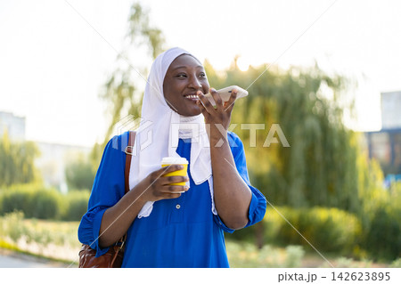 A smiling African American Muslim woman wearing a white hijab and blue dress holds a coffee cup while talking on her smartphone outdoors A smiling African American Muslim woman wearing a white hijab and blue dress holds a coffee cup while talking on her smartphone outdoors 142623895