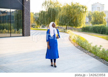 A smiling African American Muslim woman wearing a hijab and a blue dress walks outdoors holding a yellow cup 142623958