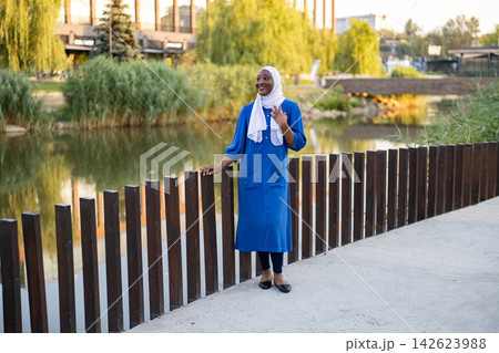 A happy African American Muslim woman wearing a blue dress and white hijab stands by a wooden fence near a body of water 142623988