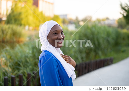 A young African American Muslim woman wearing a white hijab and blue dress smiles warmly while standing outside near lush greenery A young African American Muslim woman wearing a white hijab and blue dress smiles warmly while standing outside near lush greenery 142623989