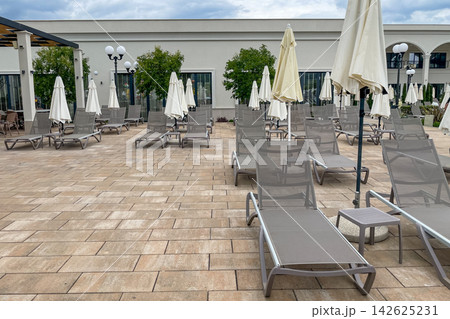 Empty sunbeds and umbrellas on pool terrace at spa resort exterior on cloudy day 142625231
