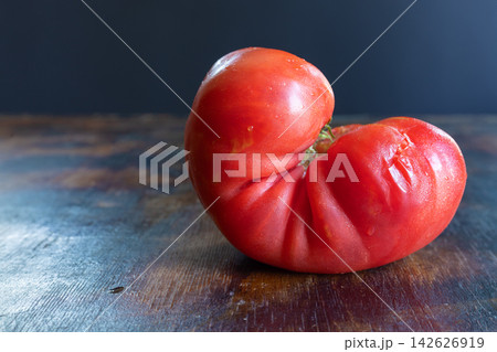Close-up still life of ugly ripe red tomato with natural folds and irregular contours, resting on rustic wooden surface. Concept of imperfect produce, natural variation, freshness, healthy eating Close-up still life of ugly ripe red tomato with natural folds and irregular contours, resting on rustic wooden surface. Concept of imperfect produce, natural variation, freshness, healthy eating 142626919