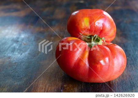 Close-up still life of ugly, naturally misshapen ripe tomato with deep folds, irregular contours, visible surface imperfections, on rustic wooden table. Concept food waste reduction, natural variation Close-up still life of ugly, naturally misshapen ripe tomato with deep folds, irregular contours, visible surface imperfections, on rustic wooden table. Concept food waste reduction, natural variation 142626920