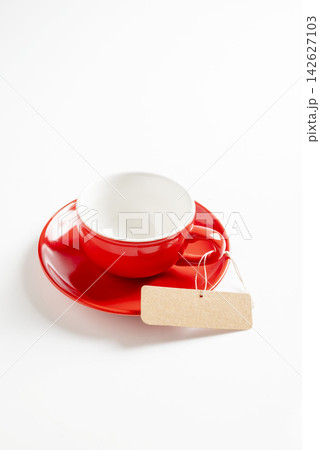 Empty red ceramic coffee cup with matching saucer and blank kraft paper tag on a white background. Template for copy space or mockup 142627103