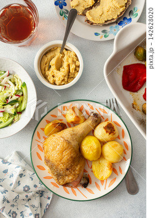 A baked chicken leg rests on a plate with potatoes. Fresh salad, roasted Brussels sprouts, and chickpea bread are arranged on the table. It looks like a wholesome meal A baked chicken leg rests on a plate with potatoes. Fresh salad, roasted Brussels sprouts, and chickpea bread are arranged on the table. It looks like a wholesome meal 142628020