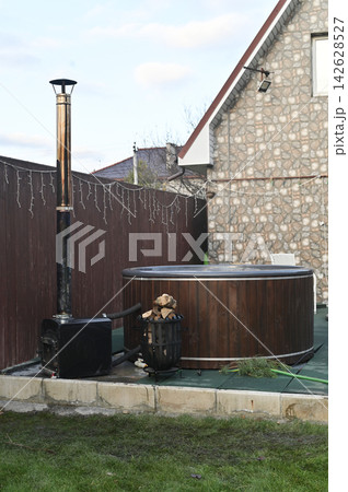 vertical photo, Metal stove positioned near round wooden hot tub on patio. Stove connected by pipe for heating water. stove creating cozy outdoor spa atmosphere, wellness lifestyle concept, 142628527