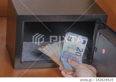 Hand holding euro banknotes depositing cash into a black safe with an open door, interior view showing a felt-lined compartment and wooden surface in the background Hand holding euro banknotes depositing cash into a black safe with an open door, interior view showing a felt-lined compartment and wooden surface in the background 142629424