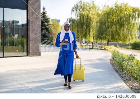 A smiling African American Muslim woman in a blue hijab and dress walks with a yellow suitcase while looking at her phone 142630609