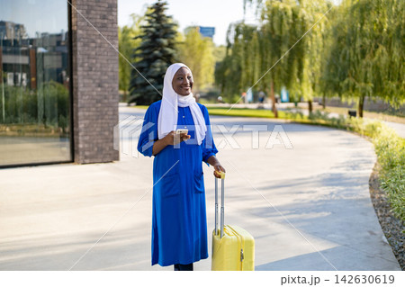 A smiling African American Muslim woman in a blue dress and white hijab stands with a yellow suitcase and phone 142630619