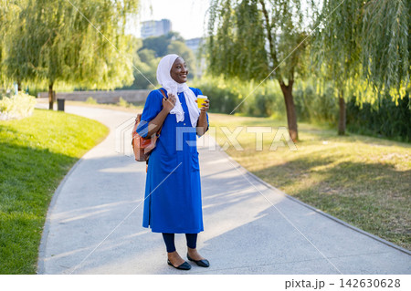 A smiling African American Muslim woman wearing a hijab and a blue dress walks in a park with a coffee cup and backpack 142630628
