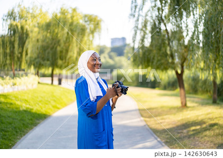 An African American Muslim woman wearing a hijab and blue dress holds a camera while walking in a park An African American Muslim woman wearing a hijab and blue dress holds a camera while walking in a park 142630643