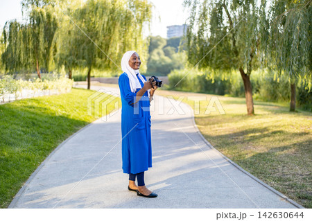 An African American Muslim woman wearing a hijab and a blue dress holds a camera while walking on a path in a park 142630644